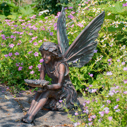 Statue of a resin fairy sitting on a stone in a garden with flowers