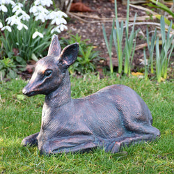 Cast aluminium statue of a small metal fawn lying on grass with plants in the background